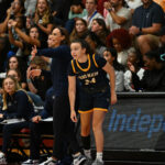 Azra Ozgul, GBC student athlete, standing by the sideline with eyes focused on the basketball game, crouching slightly in preparation for reentering the game while her coach calls instructions to the team behind her in front of the crowds in the bleachers.