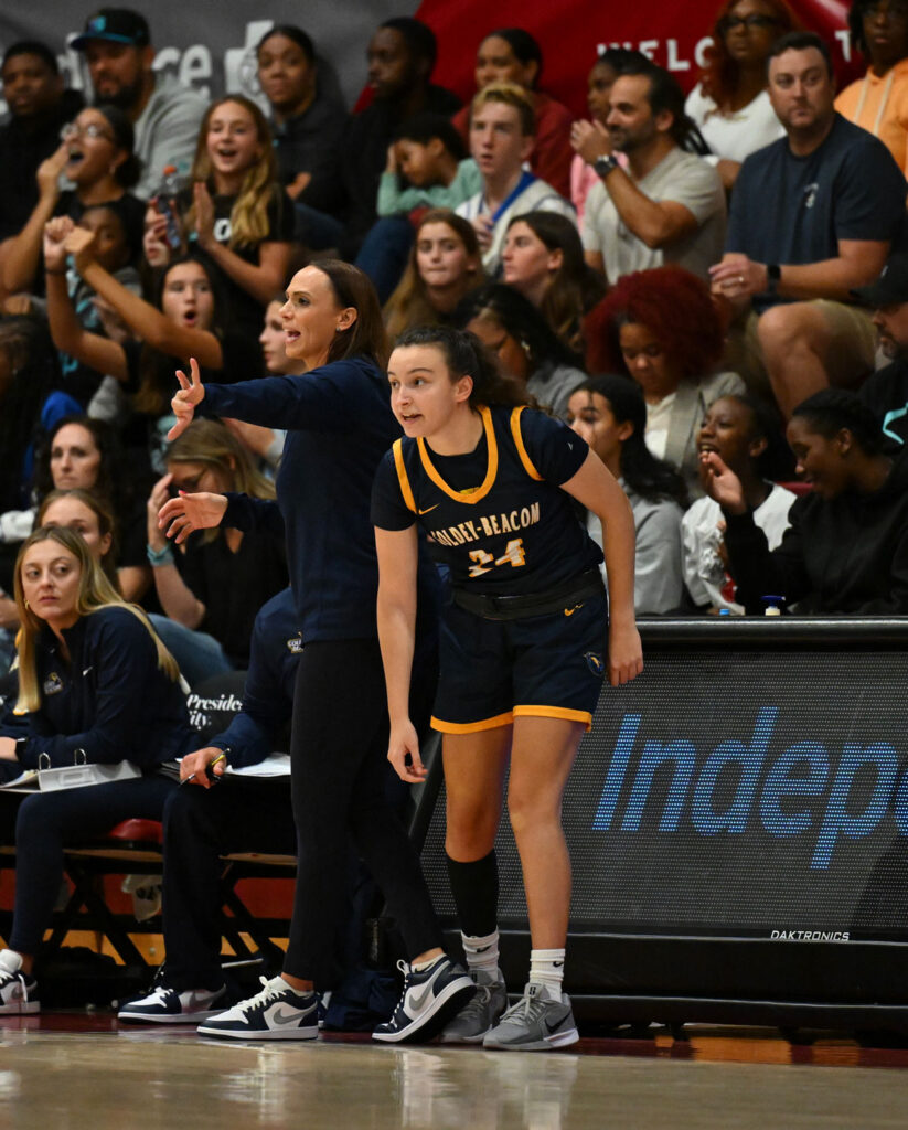 Azra Ozgul, GBC student athlete, standing by the sideline with eyes focused on the basketball game, crouching slightly in preparation for reentering the game while her coach calls instructions to the team behind her in front of the crowds in the bleachers.