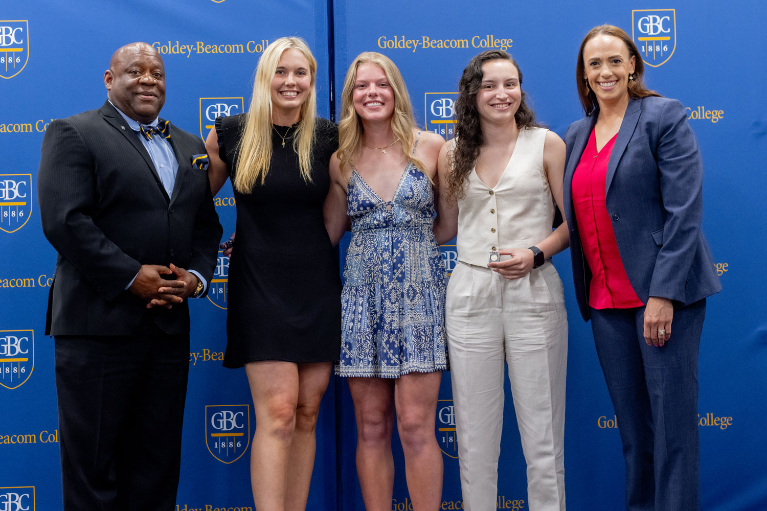 Azra Ozgul, GBC student athlete, standing with a group of students and coaches and smiling in front of the blue GBC step and repeat with the Goldey-Beacom College logo in gold and white.
