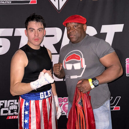 GBC student, Cesar Aguilar, poses with faculty member, Dr. Scott Glenn in front of an athletics step and repeat, wearing boxing clothes and gloves.