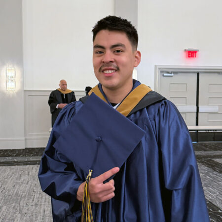 GBC graduate, Cesar Aguilar, stands with cap in hand smiling for a photo.