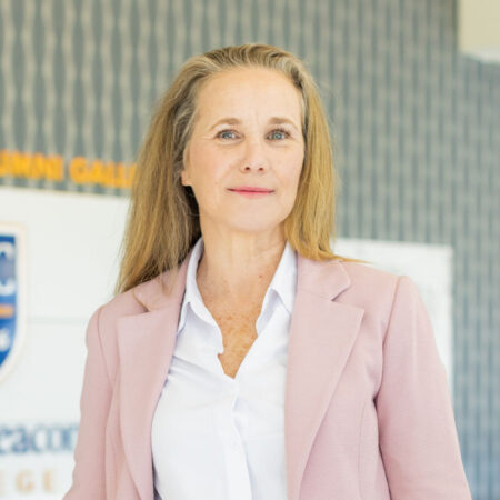 GBC alum, Cynthia Karnai, stands in front of the distinguished alumni wall wearing a pink blazer and white collared shirt.