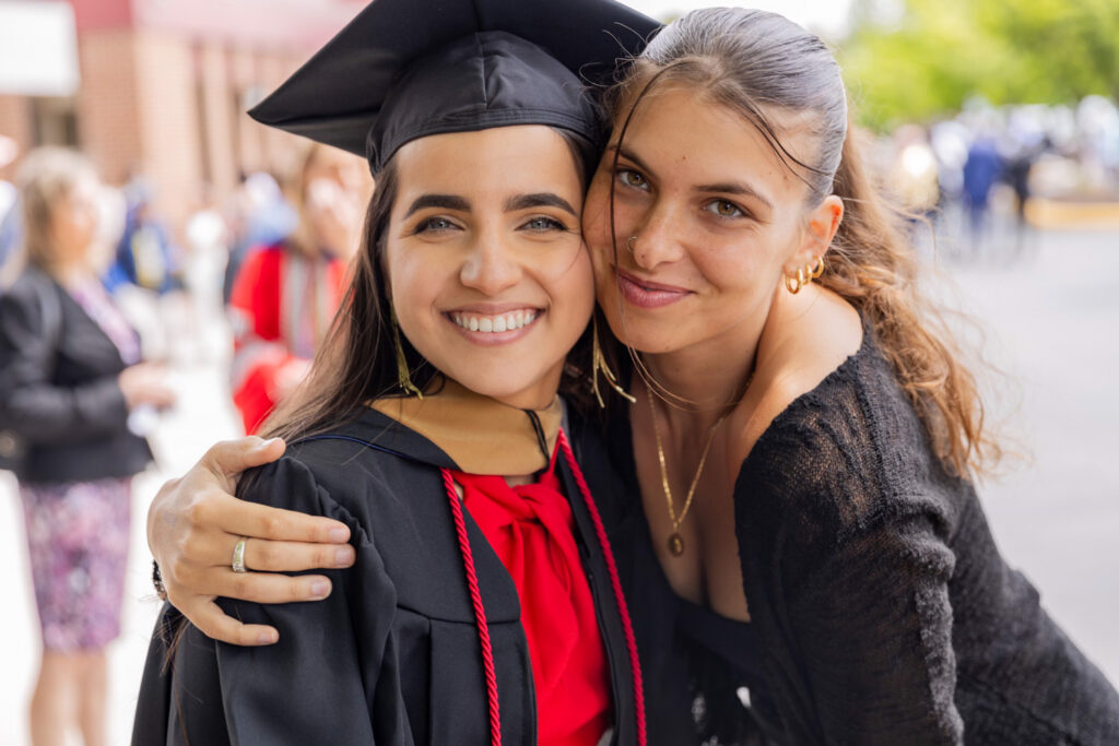 Giuliana Jaku, GBC international transfer student, stands with an arm around her friend who is wearing a cap and gown, smiling together with crowds of celebrating families behind them after graduation.