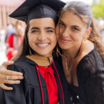Giuliana Jaku, GBC international transfer student, stands with an arm around her friend who is wearing a cap and gown, smiling together with crowds of celebrating families behind them after graduation.