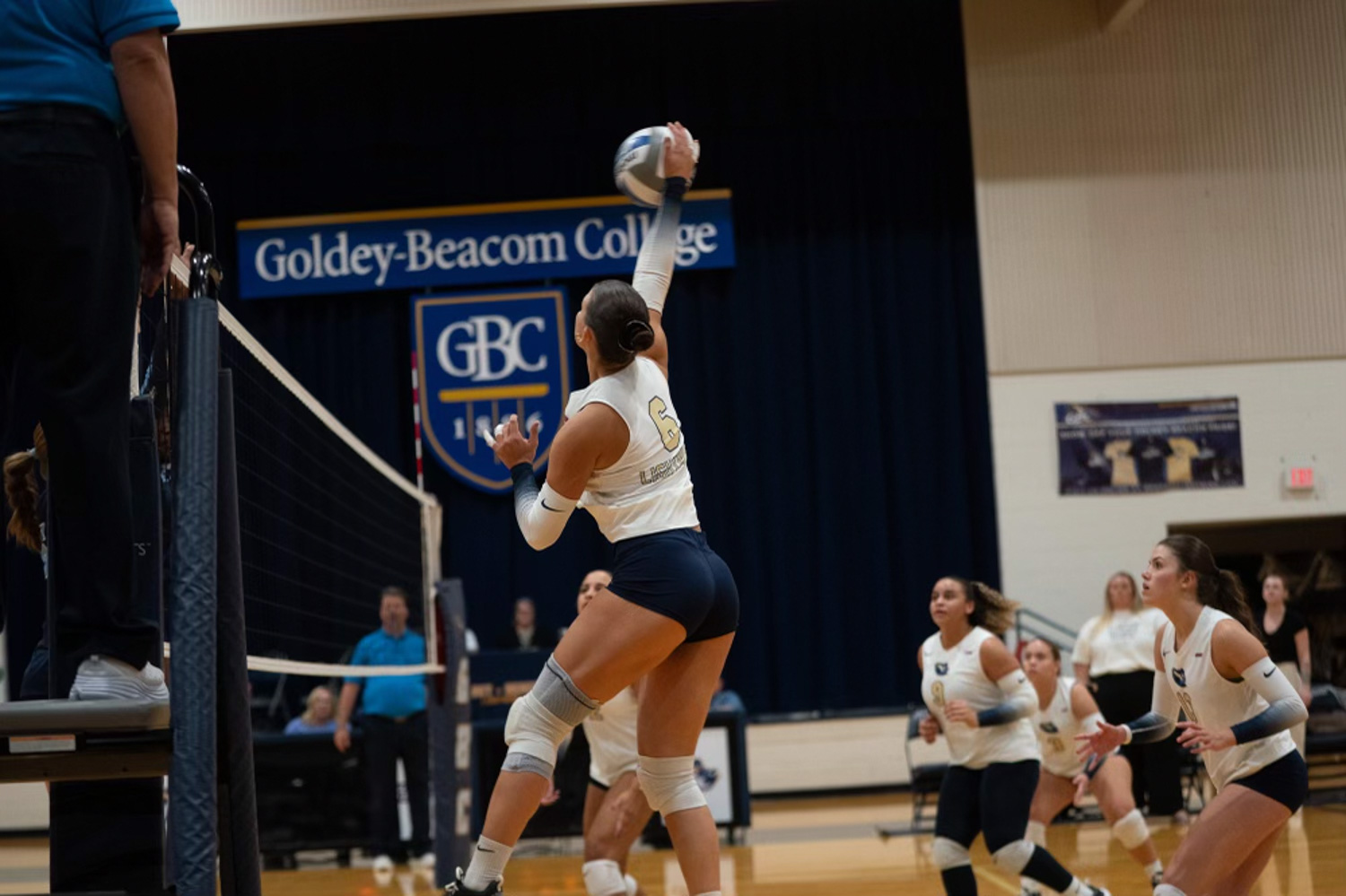 Wearing her GBC volleyball uniform, Giuliana Jaku leaps to hit a volleyball over the net while teammates watch expectantly off to the side.