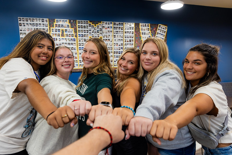 Six female students put their fists together for a group fistbump in front of a photo collage spelling out the word "Family"