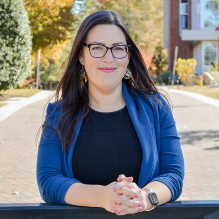 Jenn Wells, GBC Marketing Director, poses with arms leaning on a guard rail at the front of the entryway to Goldey-Beacom College's campus with green and yellow foliage visible behind her.