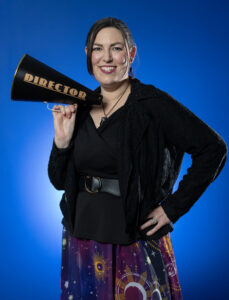 Jenn Wells, Director of Marketing at Goldey-Beacom College, stands with one hand on hip and the other holding a black bullhorn with the word "Director" in bold gold font. Jenn is smiling and wearing a very stylish black and purple ensemble.