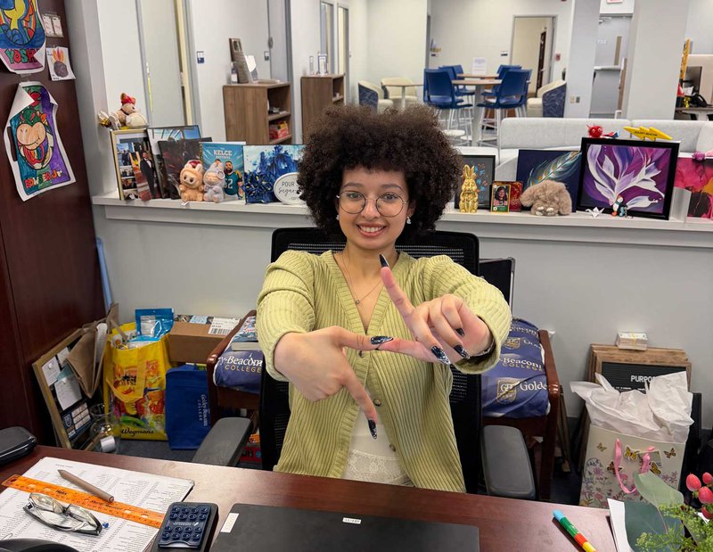 G.B.C. alumni sitting at a desk with a computer, paperwork, and hands formed into a lightning bolt shape