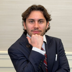 JJ Becerra poses for a headshot with his chin resting dramatically on his hand while clad in a very dapper suit.