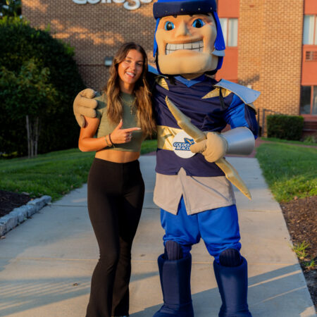 GBC student, Macy Hendin-Walker, standing next to the school mascot, Strike, each point to each other and smiling in front of the school