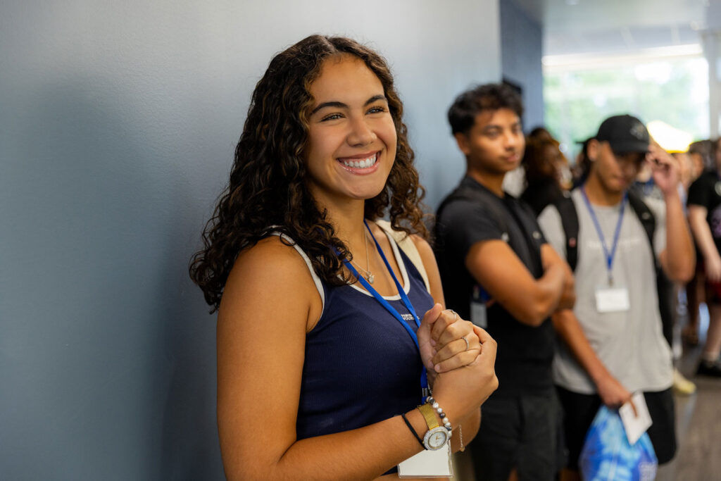 A GBC student smiles in a hallway during New Student Orientation. Students beyond her lined up further down the hallway out of focus but seemingly amused at something the photographer is saying.