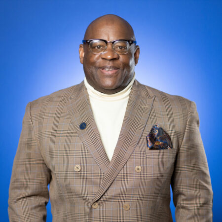 Dr. Scott Glenn, Assistant Professor of Business/NCAA Faculty Athletics Representative at Goldey-Beacom College, posing in a brown suit with white turtleneck in front of a blue background.