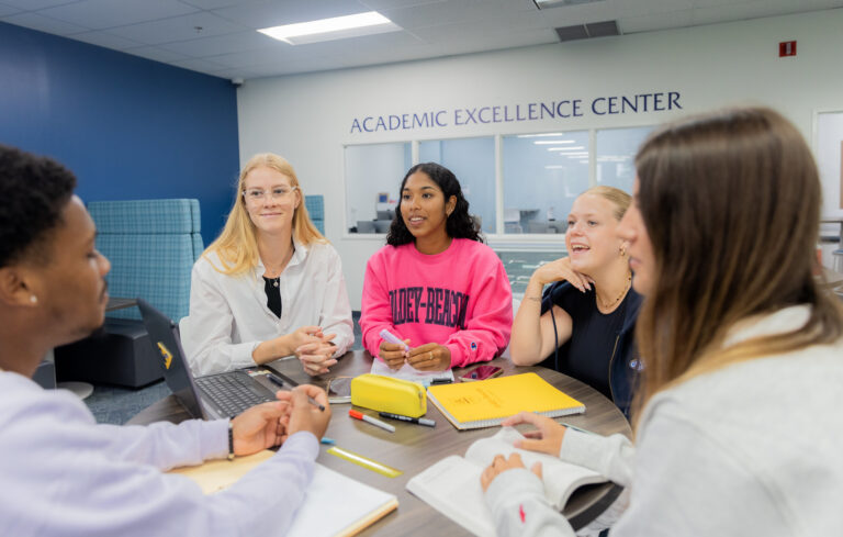 Students collaborating at a table in the Academic Excellence Center with laptops, notebooks, and study materials while discussing coursework.