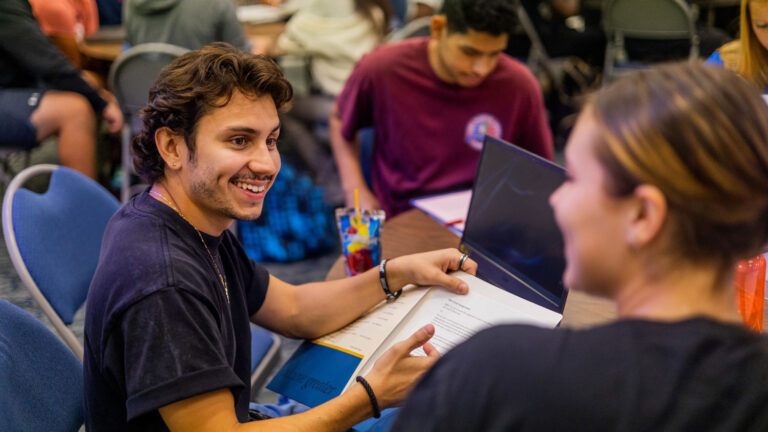 GBC student, J.J. Becerra talks to a peer advisor during new student orientation while holding a folder of GBC introductory materials in a room filled with other new students.